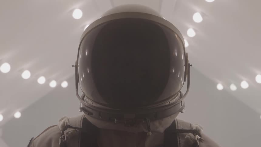 A close view of a person wearing a white and gray space suit with a dark helmet and visor standing in a sterile white corridor with many circular lights