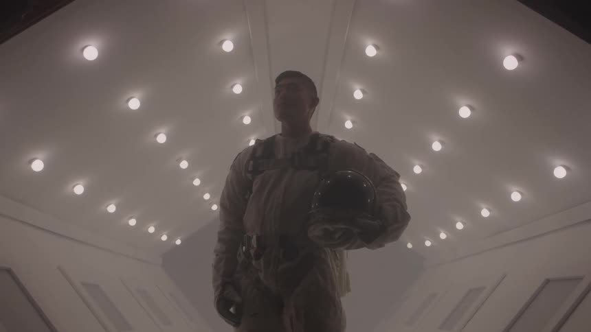 A person in an astronaut suit stands in a sterile futuristic corridor with a serious expression holding a helmet under their arm the bright lights on the ceiling create a dramatic backlit effect