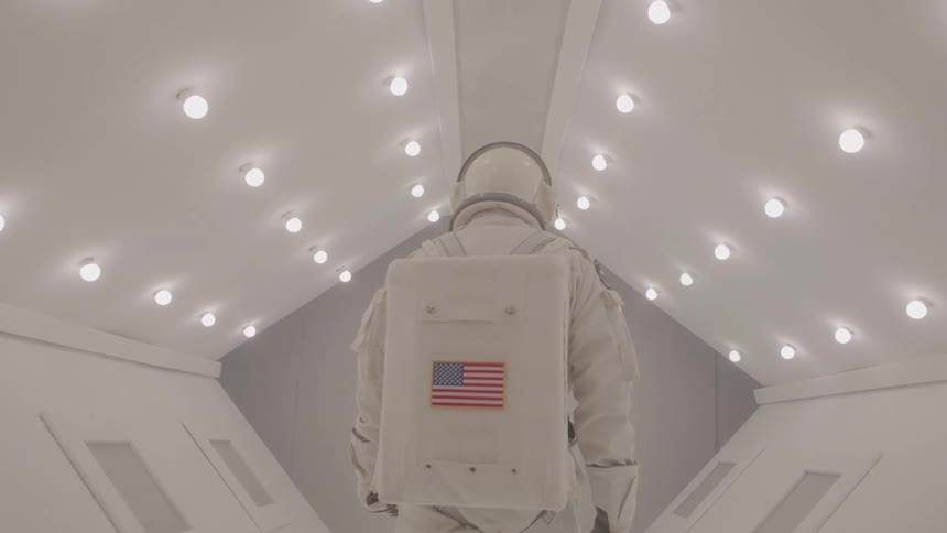 A person in a white space suit with a helmet and an american flag on the backpack walking down a sterile white corridor with many circular lights