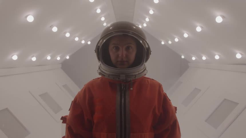 Woman wearing an orange suit and helmet standing inside an empty space shuttle illuminated by a series of small lights