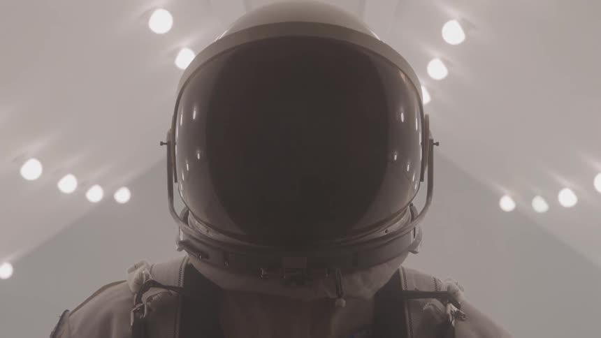 A close view of a person wearing a white and gray space suit with a dark helmet and visor standing in a sterile white corridor with many circular lights