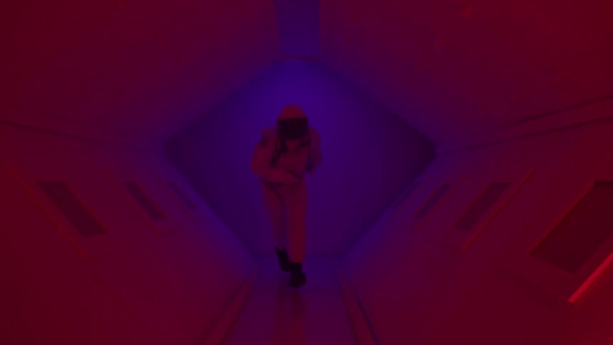 Person running inside a spaceship wearing a spacesuit and space helmet as red warning lights flicker through the dim interior
