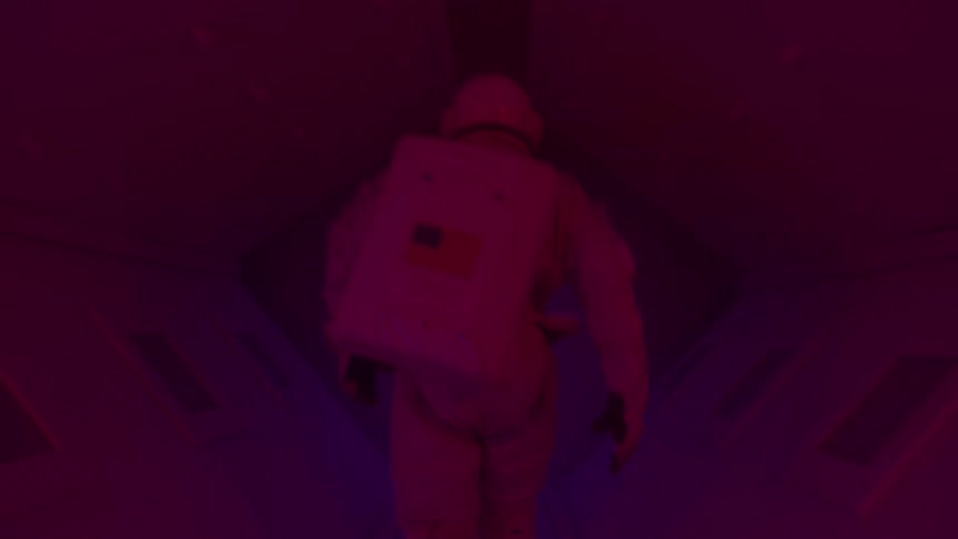 A person wearing an astronaut suit is walking away from the camera in a dark room illuminated by a mysterious red light the persons face is obscured adding a sense of suspense to the scene