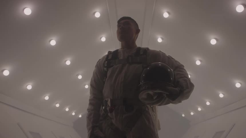 A male astronaut is standing in a brightly lit futuristic corridor holding a helmet under his arm the lighting on the ceiling creates a dramatic backlit effect and the astronaut looks determined as he stares into the distance