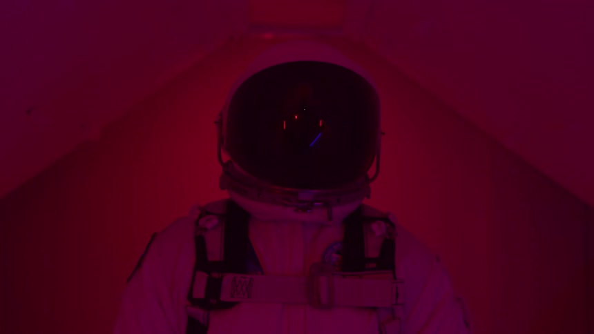 A wide view from behind shows a person in a white space suit and helmet standing in a sterile white room filled with a deep red and blue flashing lights