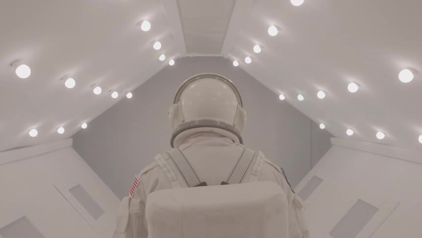 An astronaut in a white space suit stands in a sterile white hallway with overhead lights facing away from the camera