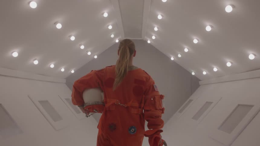 Woman wearing an orange suit holding a helmet in her left hand walking into a space station illuminated by a series of small lights