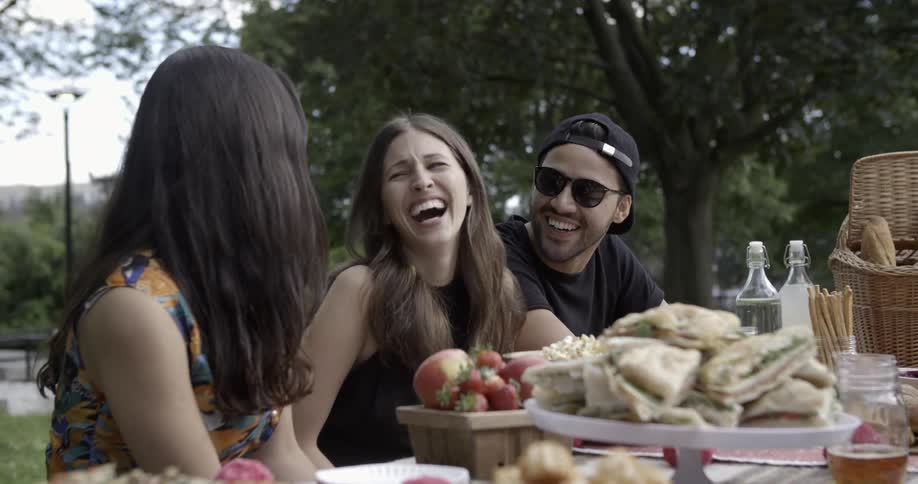 A group of people sitting at a table with food outside and laughing
