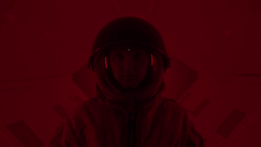 Person in an orange spacesuit with a reflective helmet in an environment lit by red and purple lights