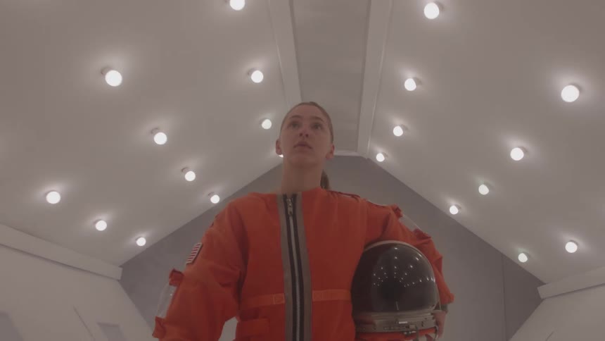 Woman in orange outfit and space helmet walking down a lit hallway
