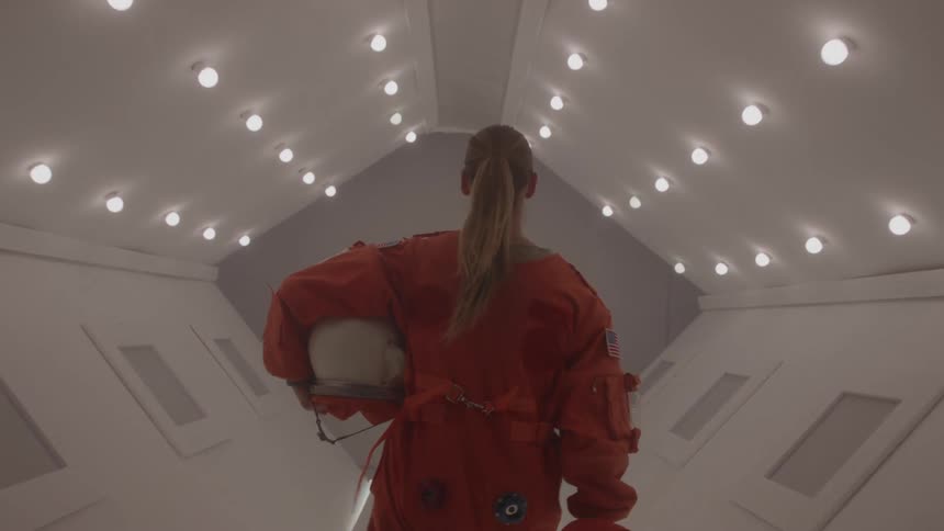 Astronaut in orange suit holds helmet while walking through illuminated corridor