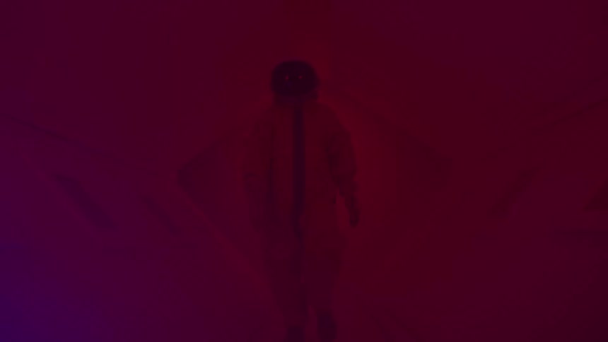 A silhouetted person wearing a red jumpsuit and a helmet walks through a foggy room with changing red and purple lighting