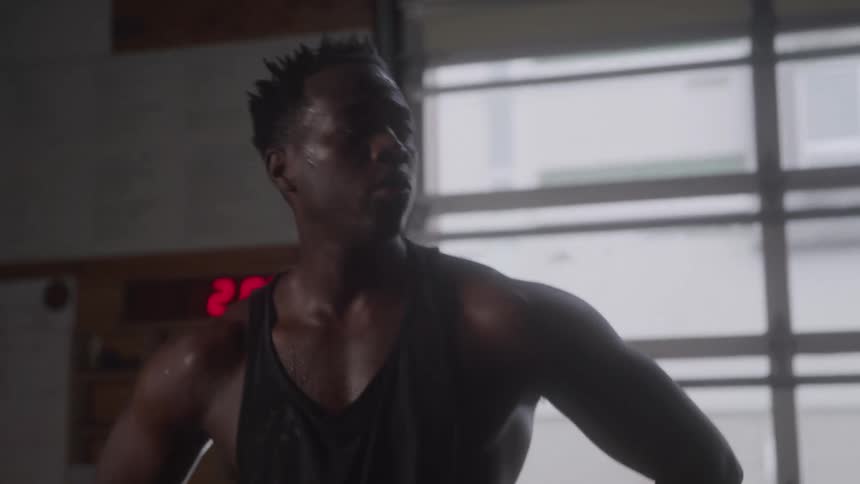 A close view of a sweating man in a black tank top breathing heavily and looking up to the side in a dimly lit gym with a red digital clock