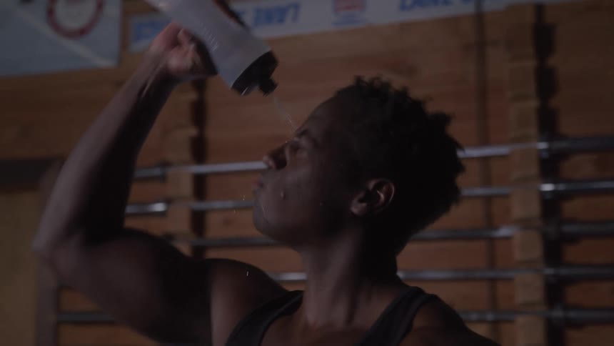 A close view of a man in a black tank top drinking from a clear water bottle in a gym with wooden paneling on the wall and a dark moody atmosphere