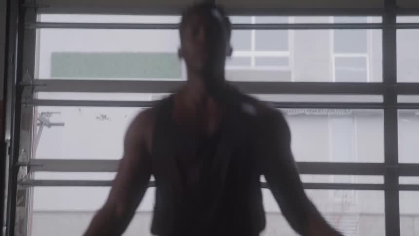 A man wearing a black tank top and shorts is jumping rope in a gym with large windows in the background