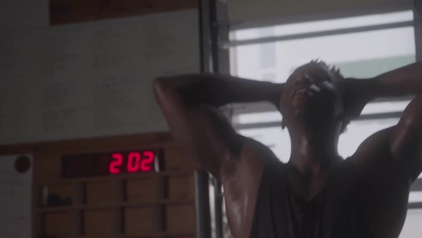A sweaty muscular man in a tank top runs his hands through his hair looking exhausted