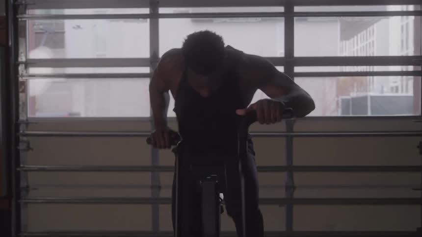 A man in a black tank top leans forward on an air bike straining with exhaustion and focus