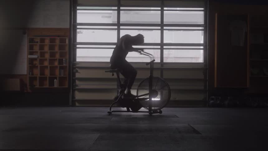 Silhouetted against a large window a person vigorously rides a fan bike in a dark gym