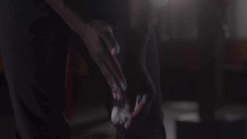 A close view shows a mans chalking his hands with outstretched fingers as he stands in a dark gym with a bright light in the background
