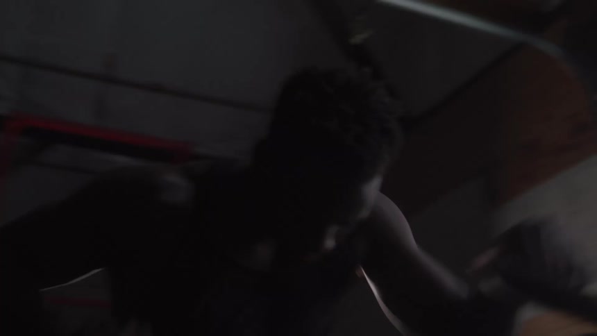 A man wearing a black tank top and shorts pedals an exercise bike with intense effort in a dimly lit gym with large windows and a bright building in the background
