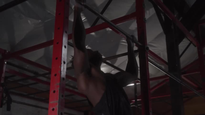 A lowangle shot shows a man doing pullups on a metal bar inside a gym the mans back muscles are flexed as he lifts his body up and down and the gym equipment and rafters are visible in the background