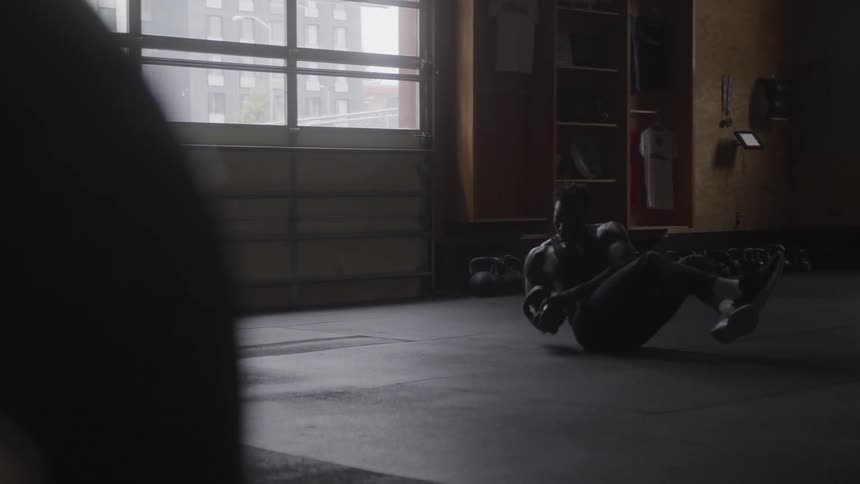 A person wearing dark workout clothes is doing an ab exercise on a dark gym floor in front of a window with a shadowy foreground and various gym equipment in the background