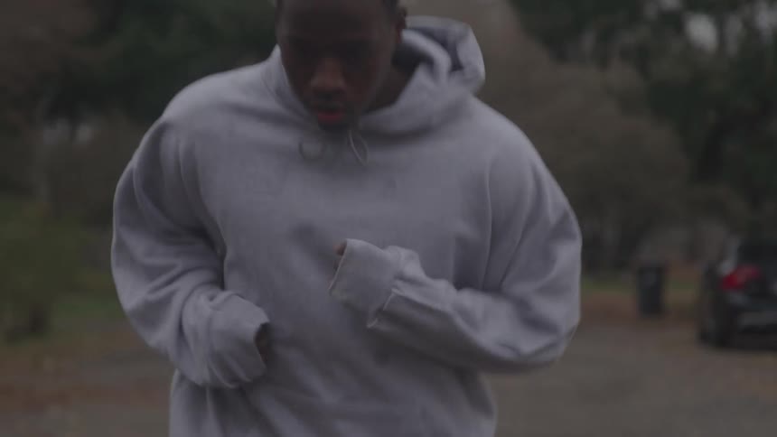 A close view of a man in a gray hoodie jogging slowly on a street on a cloudy day with trees and a car visible in the background