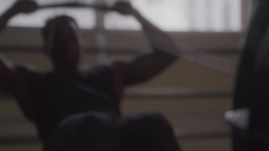 In the first video a person is shown working out on a rowing machine in a gym with their silhouette visible against a bright window the second video features a figure in a white spacesuit walking down a narrow dimly lit hallway that is cast in an unse