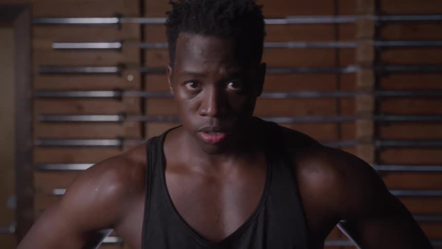 A man wearing a black tank top stands with his hands on his hips looking down while sweating heavily in a dim gym with metal barbells on a wooden wall