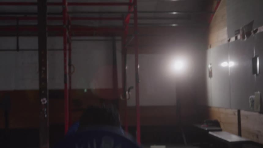 A man in a dark tank top is lifting a heavy barbell with blue and white plates in a dimly lit gym with white lockers and benches in the background