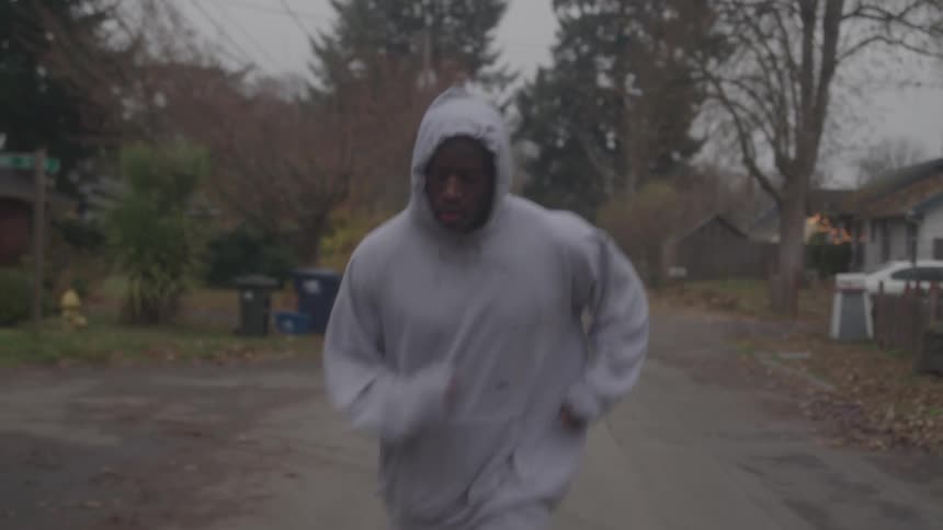 A man in a gray hoodie jogs down a quiet neighborhood street on a cloudy day looking determined