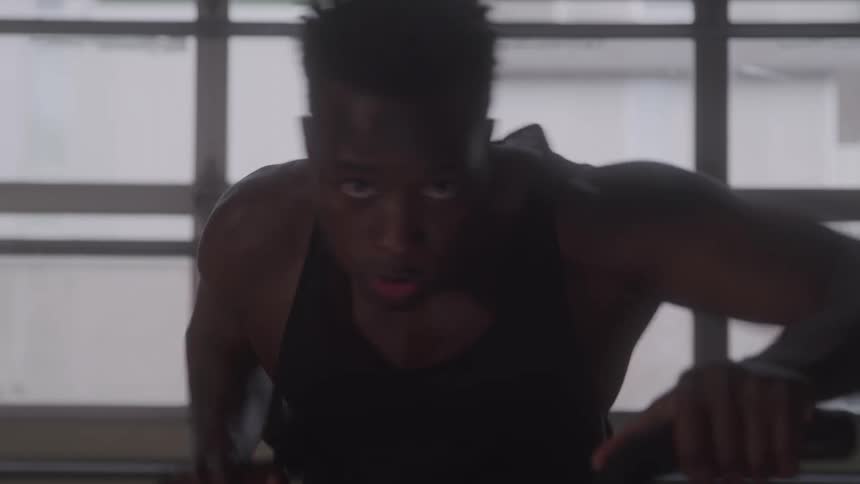 A silhouette of a muscular man wearing a tank top is doing an exercise on a machine in a brightly lit gym with large windows