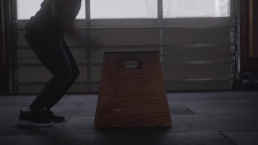 A person is jumping onto a wooden plyometric box in a dimly lit gym