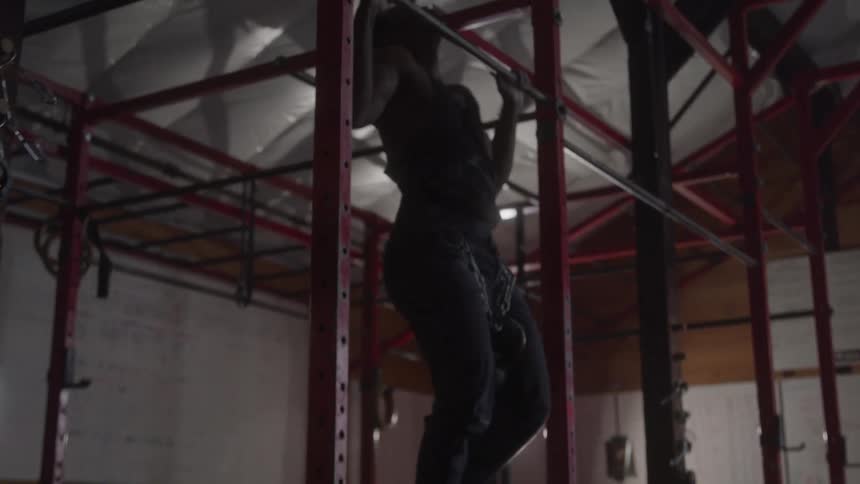 A muscular man does pull ups on a red bar in a dimly lit gym