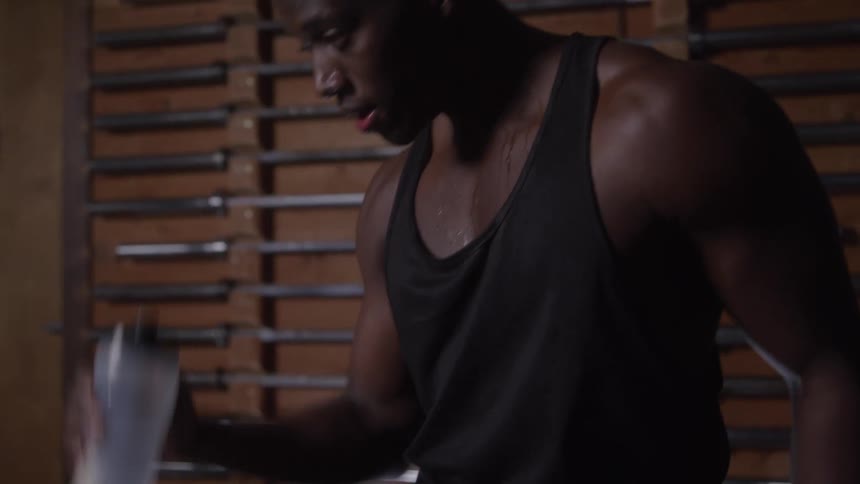 A close view of a sweating man in a black tank top breathing heavily in a dim gym with metal barbells racked on a wooden wall in the background