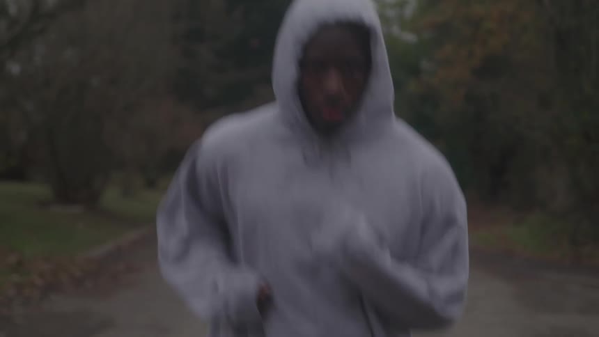 A close view of a man in a gray hoodie jogging on a street with a moody atmosphere and bare trees in the background
