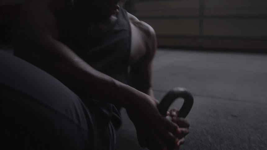 A man in a dark tank top is swinging a black kettlebell in a dimly lit gym with large windows and a timer in the background