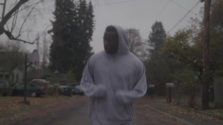A man wearing a gray hoodie and sweatpants jogs on a quiet treelined suburban street on a cloudy day