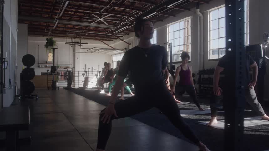 A group of men and women of various ages are doing a yoga pose with outstretched arms and bent knees in a sunlit gym