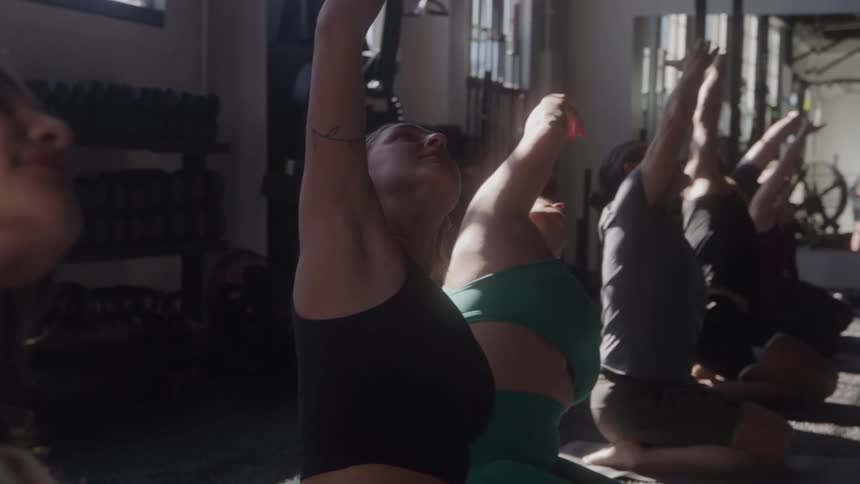 A side view of a group of men and women of various ages doing a yoga pose with their arms raised and heads tilted back in a gym with sunlight coming through the windows