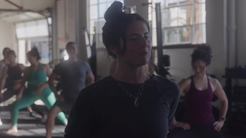 A female fitness instructor with her hair in a bun leads a diverse group of people in a workout class inside a sunlit gym