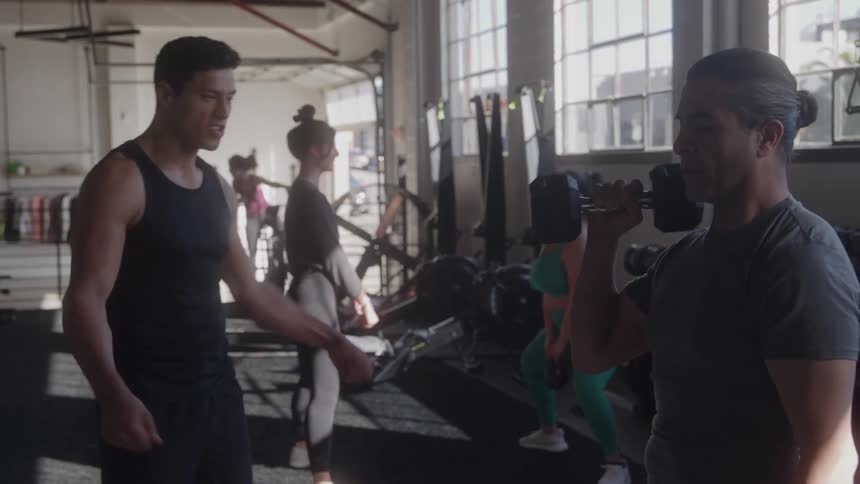 The video shows two men talking in a gym with one of them demonstrating a dumbbell curl other people are visible in the background also exercising