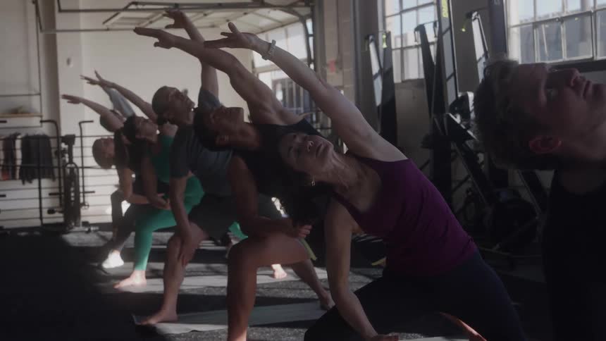 A diverse group of men and women are doing yoga in a bright modern studio with large windows and various fitness equipment in the background