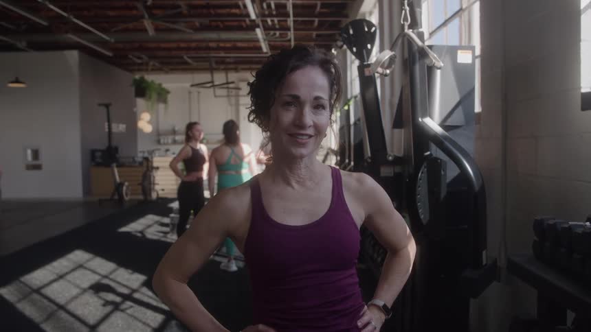 An athletic woman on a gym wearing an active tank top smiling with her hands on her hips while two women talk behind her