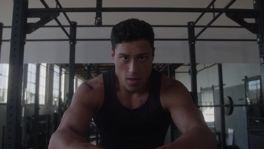 A fit man wearing a black tank top is resting in a gym with a serious expression and fitness equipment in the background