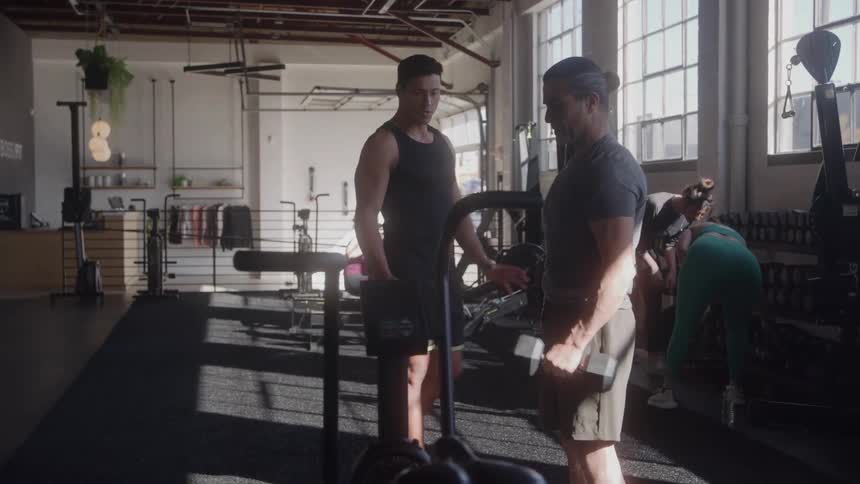 A man is coaching a male client who is lifting a dumbbell in a large gym with bright windows and other people exercising