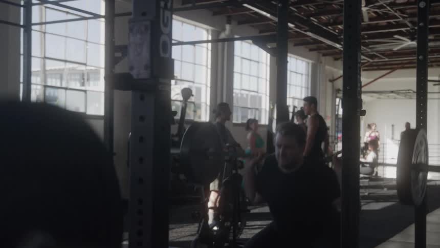 A man in a gym lifts weights while people behind him exercise in other ways