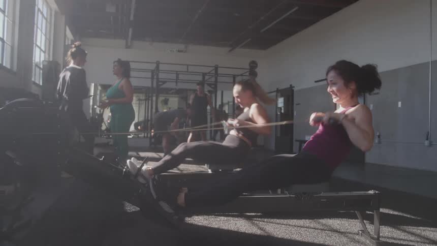 The video shows a gym scene where a group of people are exercising the main focus is on two women who are actively using rowing machines