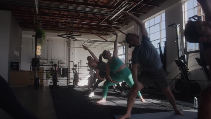 The video shows a group of people doing yoga in a gym they are performing a yoga pose possibly a variation of the extended side angle pose or triangle pose with their arms raised and bodies extended