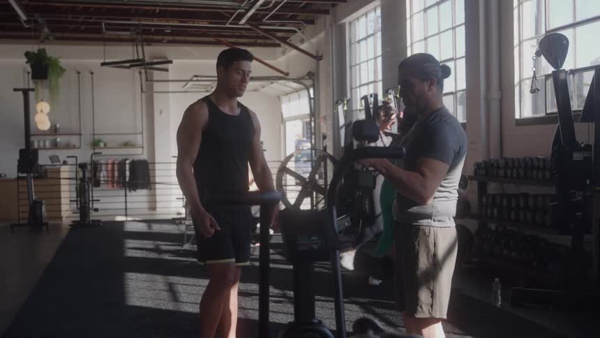 Man observes another doing bicep training in a gym lit by natural light